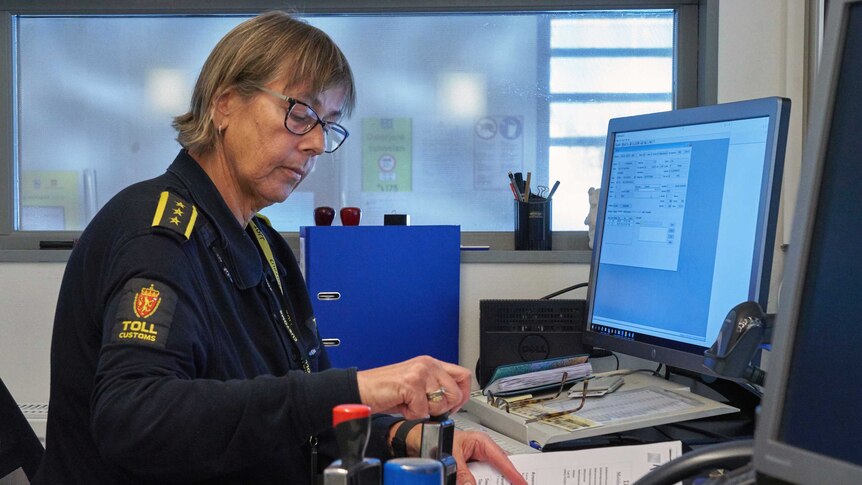 A blond woman in Norwegian 'toll customs' uniform stamps a paper form at her desk looking out into icy-blue light.