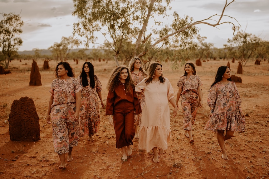 Seven Indigenous women walk through a desert landscape wearing pink and deep red patterned clothing