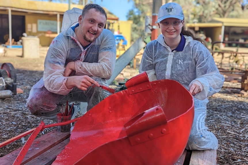 A young white man, Bryce, and Paris a young white woman with glasses and grey hat in coveralls paint a tractor seat red. 