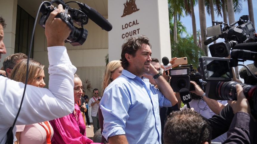 A white man in a blue collared shirt, surrounded by reporters and cameras. A sign behind him says 'Local Court'.
