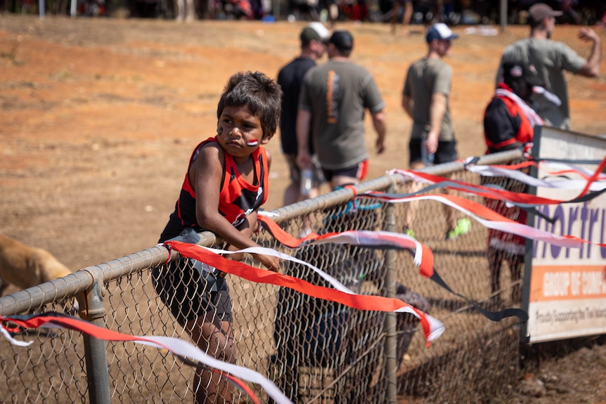A young Indigenous boy wearing a sports uniform watches an AFL game from behind a fence.