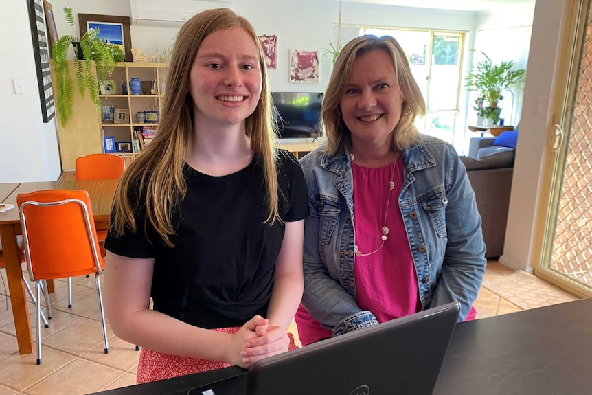 A young woman with blonde hair and an older woman stand behind a laptop computer