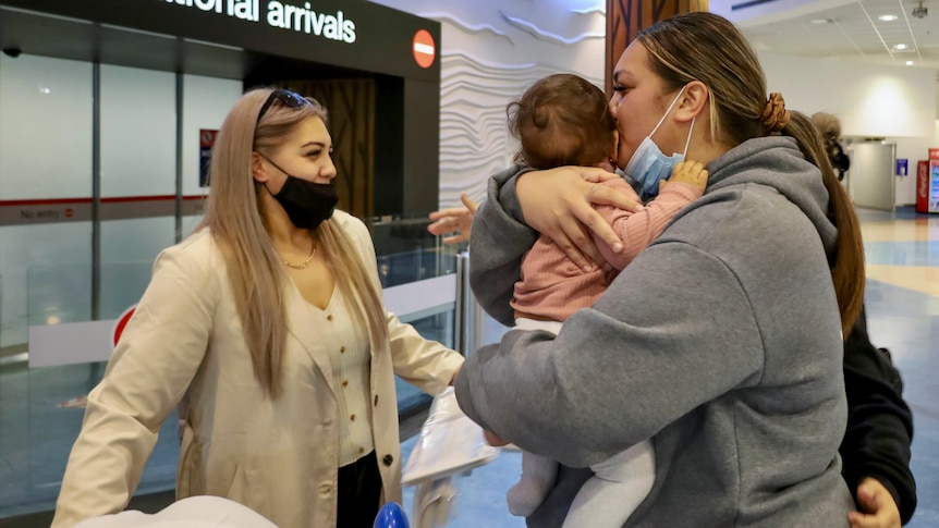 Blonde woman standing outside international arrivals hall at the airport smiling as another woman embraces a baby.
