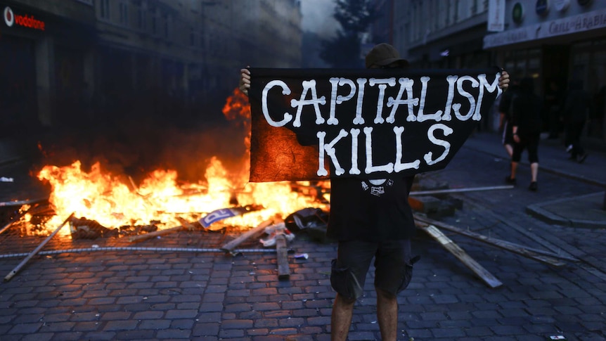 A protester wearing black holds a sign saying "capitalism kills" in front of a fire.