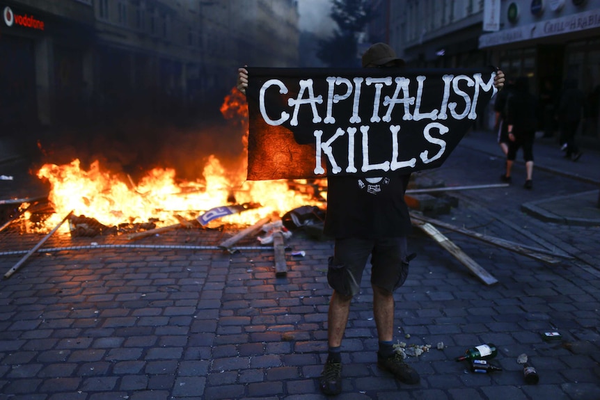 A protester wearing black holds a sign saying "capitalism kills" in front of a fire.