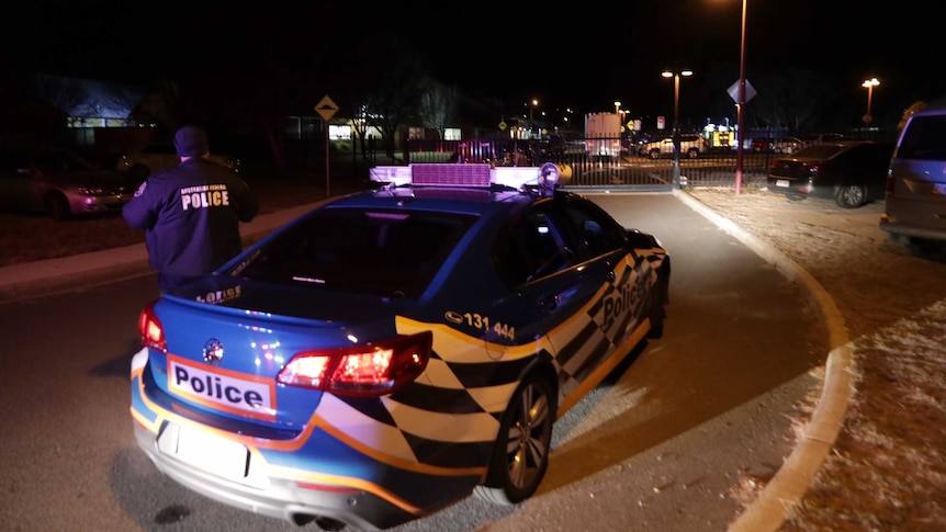 A police officer stands by a police car outside St Clare of Assisi Primary School car park.