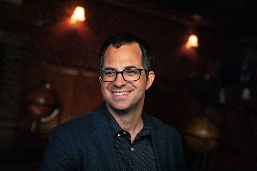 A man with dark rimmed glasses, wearing a dark shirt and jacket, smiles in a low-lit room 