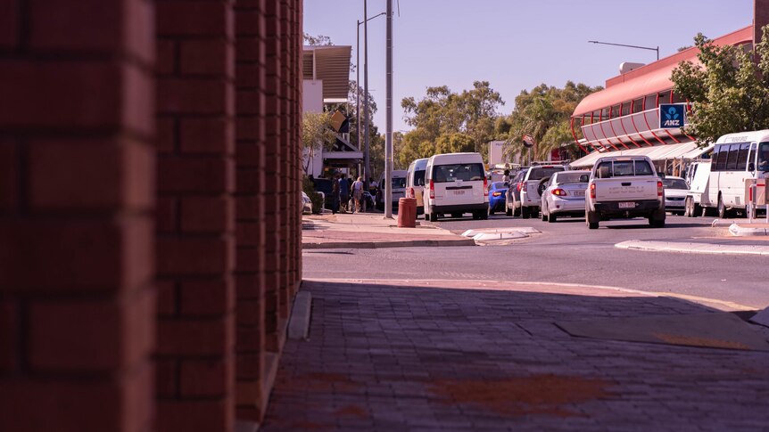 Close-up of brick wall and paved sidewalk as cars stop in traffic in front of shops and  signs of Anz and CWA bank.