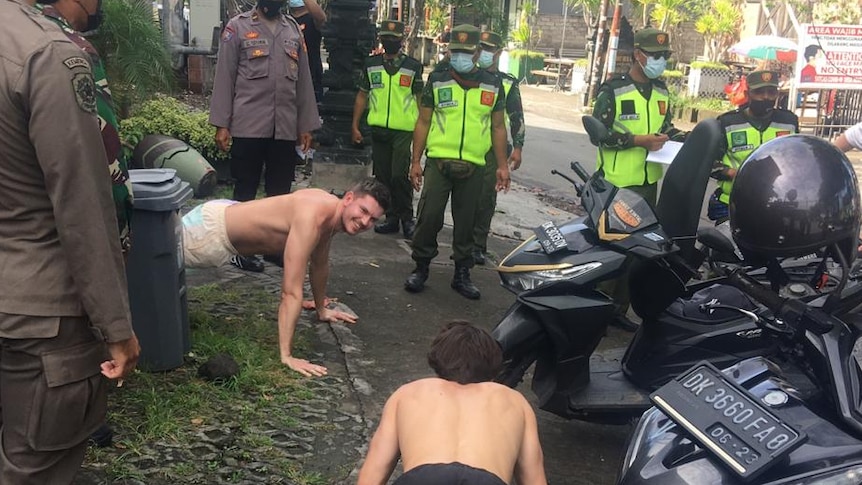 Dua orang sedang melakukan push-up di jalan-jalan Bali yang dikelilingi oleh petugas polisi.