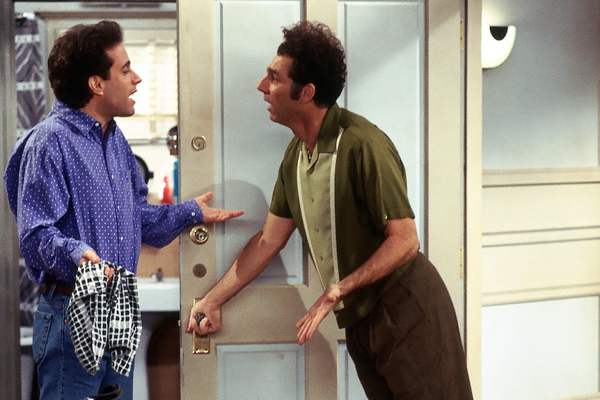 Kramer leaning on Jerry's door holding the door handle while Jerry faces him shrugging his shoulders, holding a tea towel