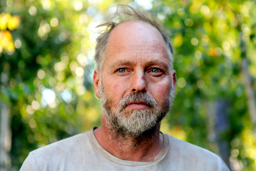 Man with thinning grey hair and a greyish beard looks into distance with trees behind