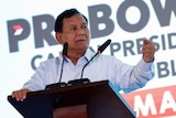 A close up of a man wearing a blue button up shirt standing in front of a podium delivering a speech.