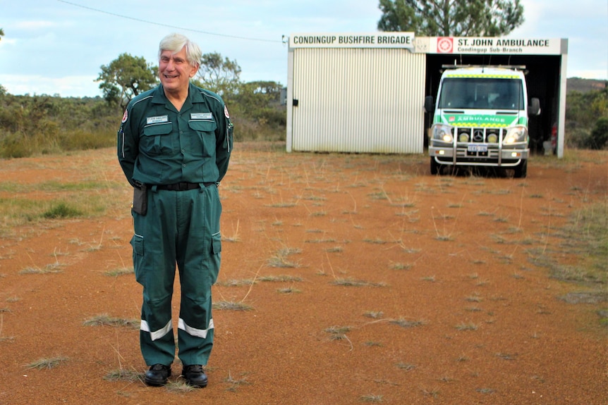 He stands in red dirt in his uniform, the ambulance is behind in a tiny tin shed