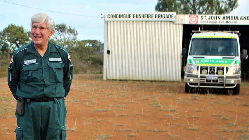 A smiling grey-haired man stands in red dirt in his uniform, the ambulance is behind in a tiny tin shed.