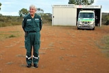 A smiling grey-haired man stands in red dirt in his uniform, the ambulance is behind in a tiny tin shed.