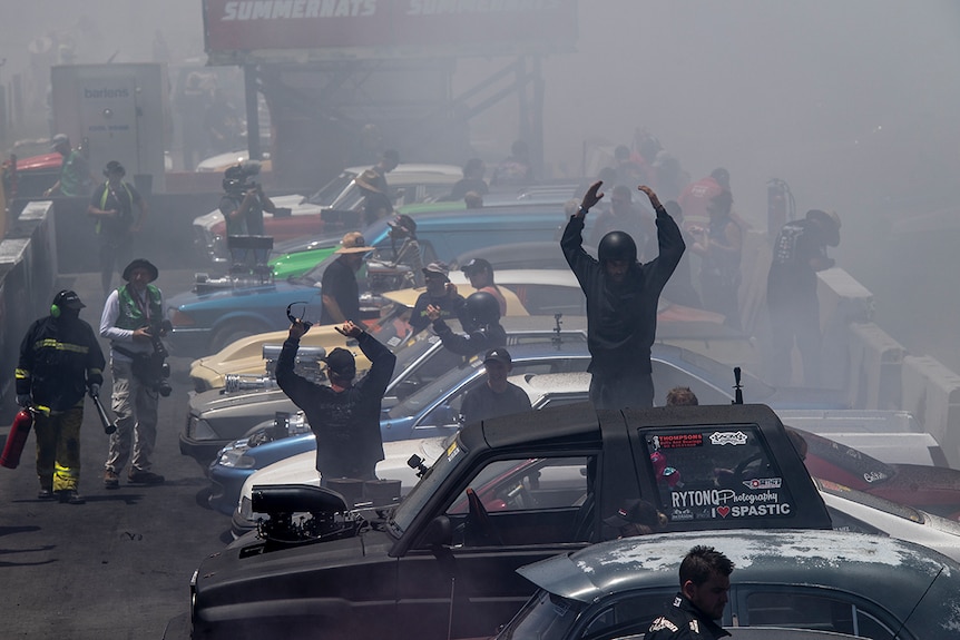 A line of drivers stand next to their cars, with one standing on his car, while surrounded by smoky air at Summernats car show