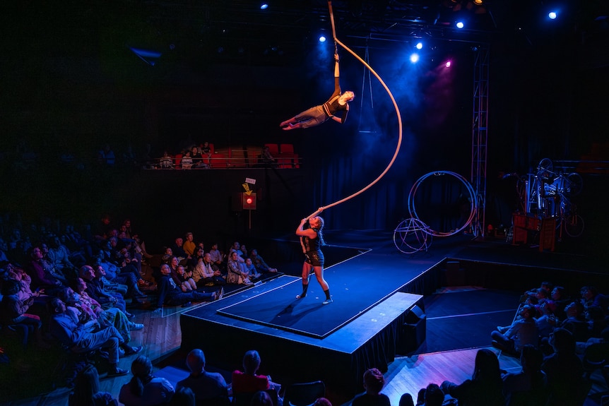 One circus performer on a darkened stage holds a rope while another holds in with one hand and swings in the air