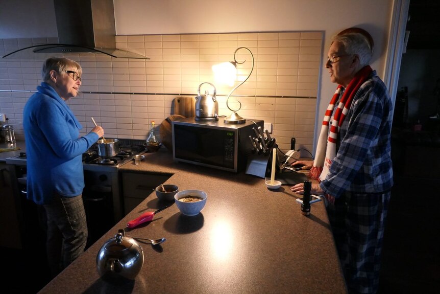 A woman stirs a pot on the stove while talking to a man in a gloomy kitchen.