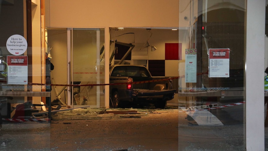 The car is lodged in the back wall of the bank, surrounded by broken glass and emergency tape.