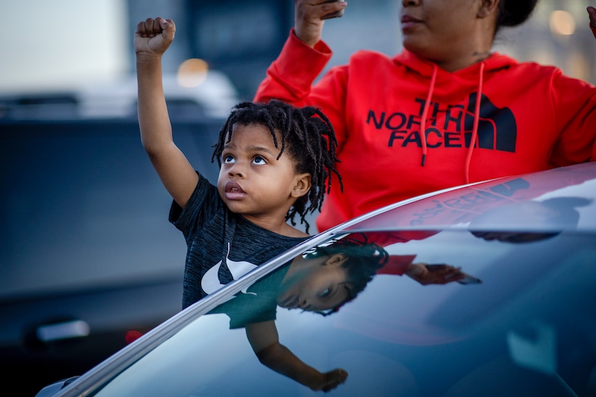 A small black boy raises his fist next to a car