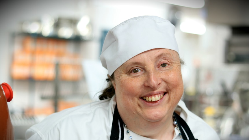 A woman smiles at the camera wearing a chef hat and coat.