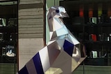 A giant pigeon made out of steel stands in a mall as people stop and look at it.