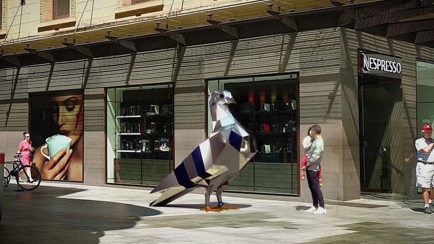 A giant pigeon made out of steel stands in a mall as people stop and look at it.