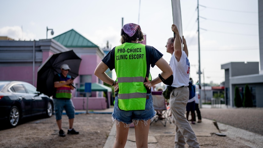 A woman wearing a hi-vis vest with PRO-CHOICE CLINIC ESCORT stands with her hands on hips, facing protesters with signs