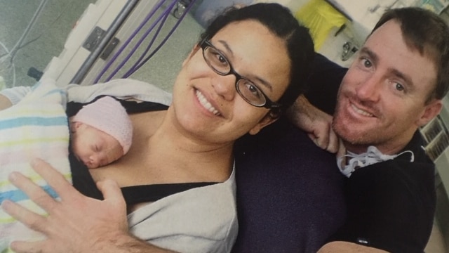 A woman holding her baby and her partner standing behind her in hospital.