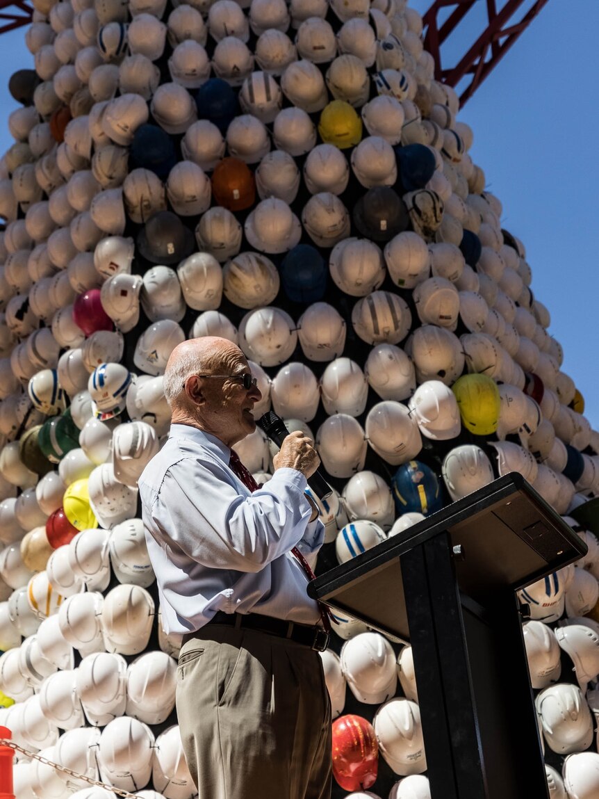 A man speaking into a microphone in front of a Christmas tree made of safety helmets.