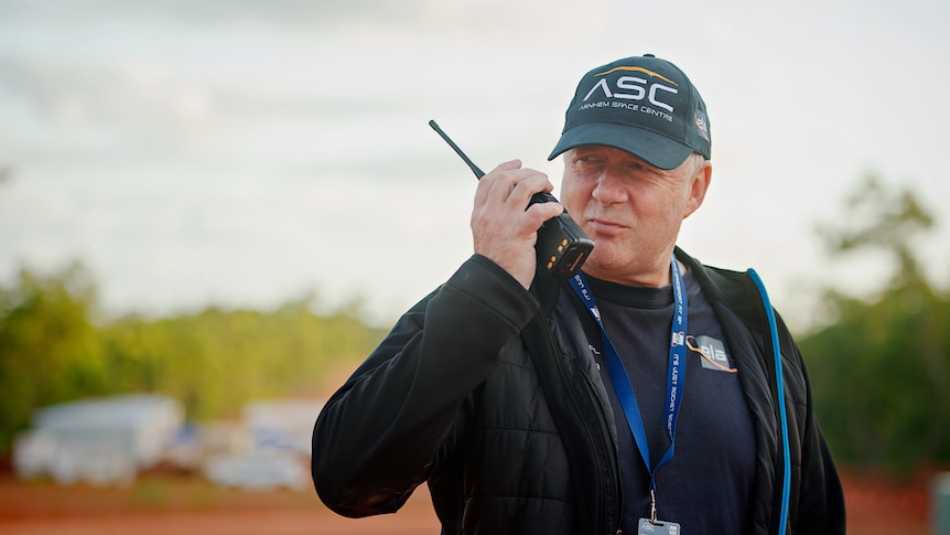 A man wearing a cap and a lanyard holds a walkie talkie to his mouth. 