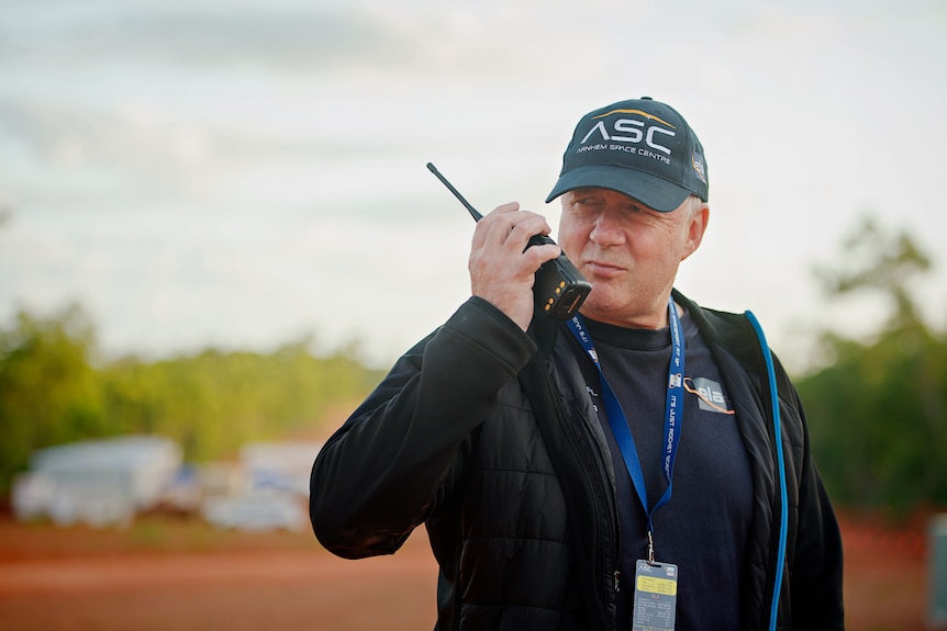 A man wearing a cap and a lanyard holds a walkie talkie to his mouth. 
