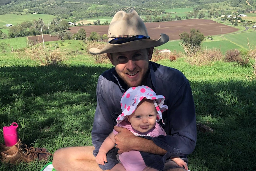 Elly's husband siting on the ground on a colourful picnic rug holding their baby daughter the farm which is looking very green