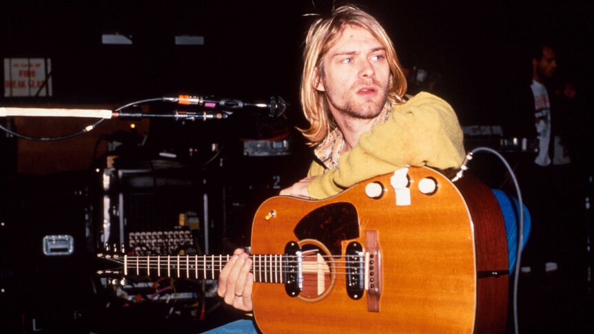 Kurt Cobain of Nirvana sits and rests a guitar on his lap.