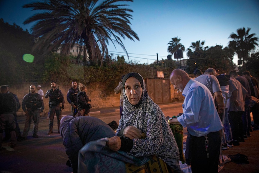 A woman in veil holds a prayer mat in a street at dusk as soldiers look on