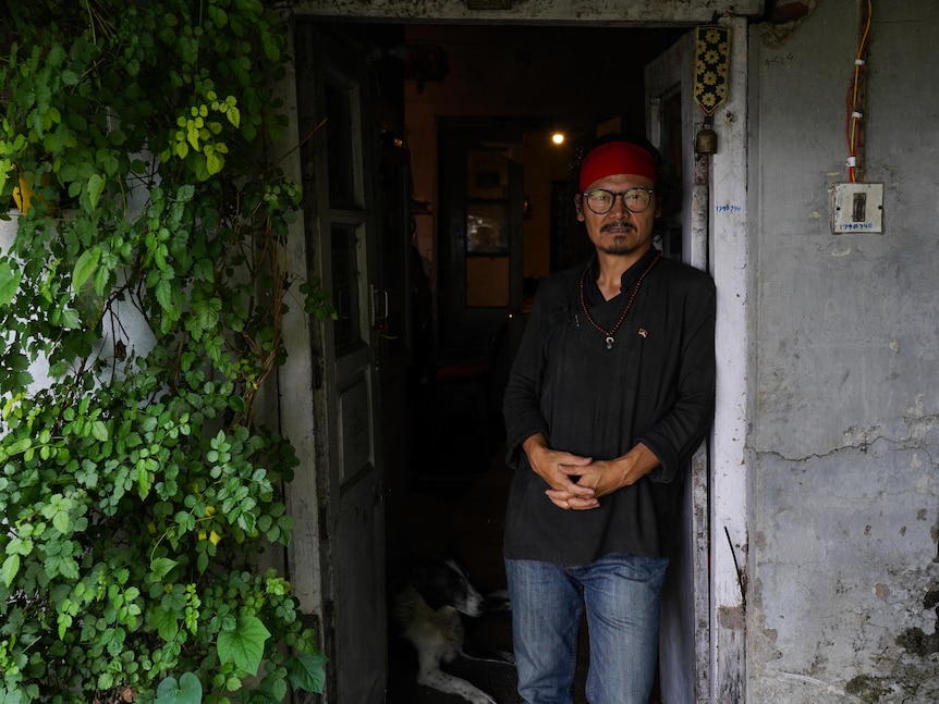 A man in a black shirt and red bandana leans against the open door of a house. Green foliage climbs the outside