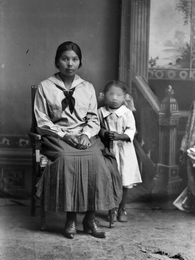 A black-and-white image showing a mother in old-fashioned clothing next to a little girl, whose face is blurred out.