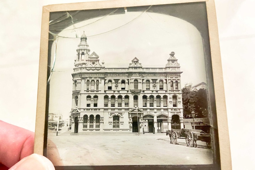 A hand holds up a glass slide photograph of a building in Brisbane in the 1800s.