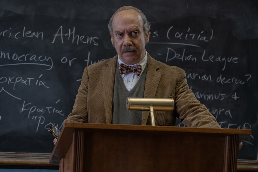 A film still of Paul Giamatti, standing behind a lectern in front of a blackboard. He has an angry expression.