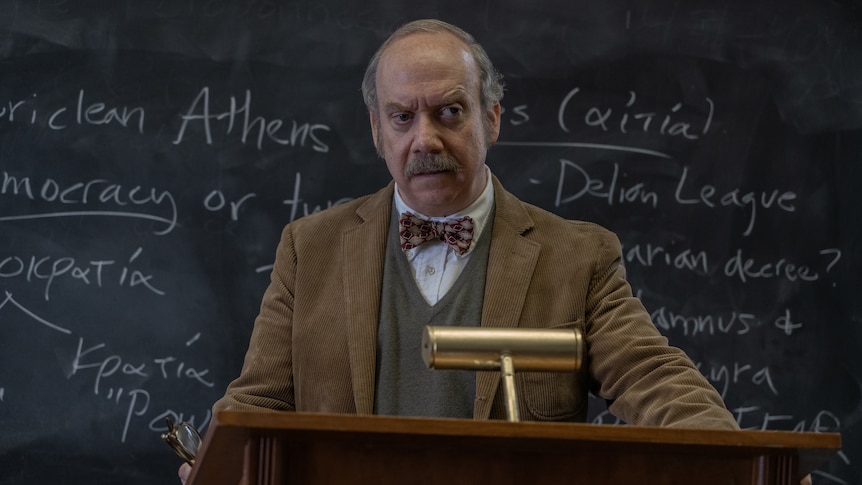 A film still of Paul Giamatti, standing behind a lectern in front of a blackboard. He has an angry expression.