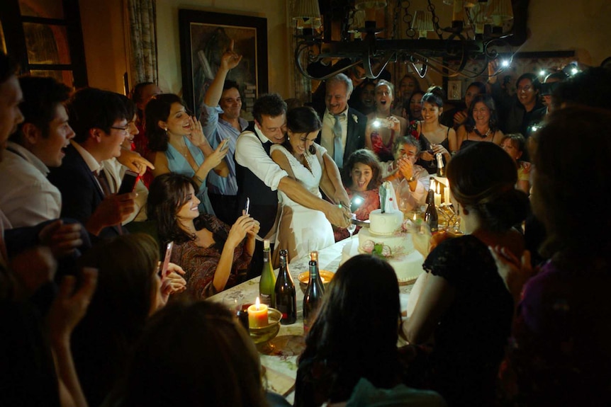 The cast, in formal wear, gather around the actors playing a bride and groom as they cut a wedding cake together.