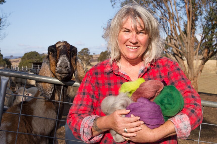 A woman with blonde hair stands with a goat in a paddock. She holds vibrant dyed wool.