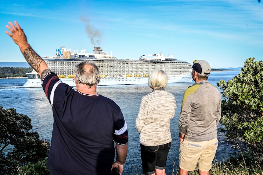 People on a headland wave to a large cruise ship seen in the distance.
