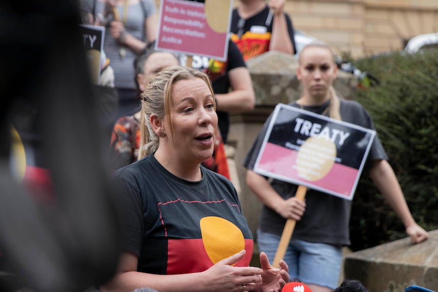 A woman with blond hair in a black, orange and red t-shirt speaks to a gathering with placards behind her