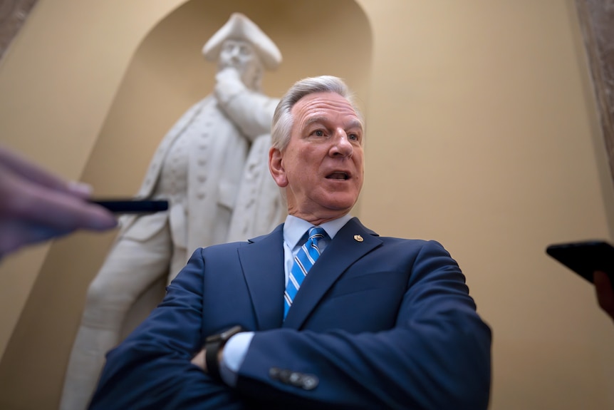 Tommy Tuberville stands with his arms folded and speaks as phones are held in front of him.