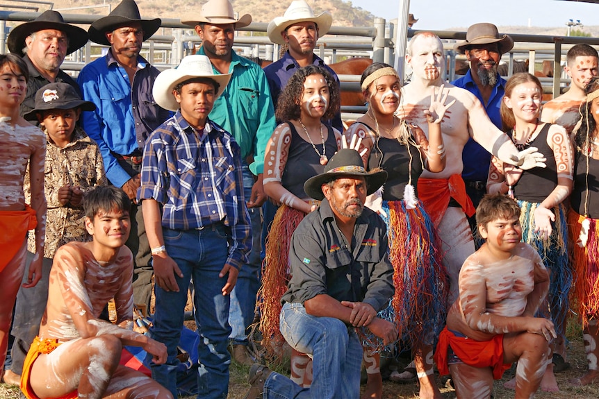 A group of Indigenous rodeo riders and dancers, all wearing colorful shirts and traditional garments pose for a group photo 