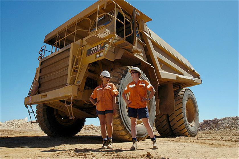 Two people in high-vis orange shirts and navy shorts stand in front of a massive mining truck.