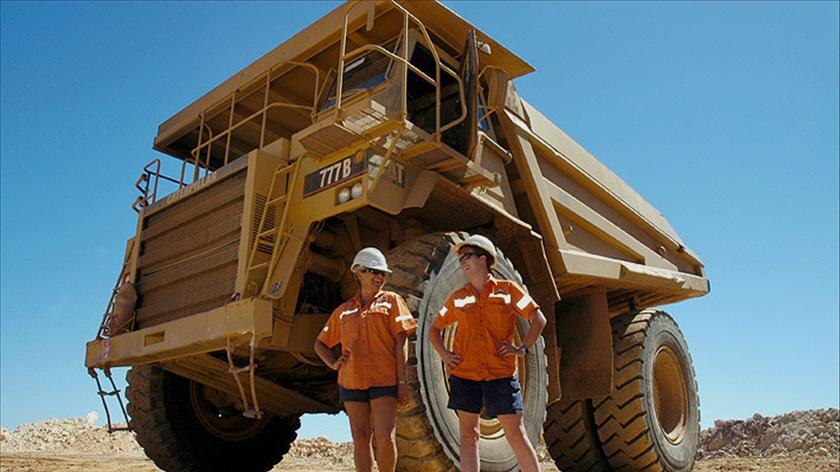 Two people in high-vis orange shirts and navy shorts stand in front of a massive mining truck.