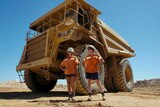 Two people in high-vis orange shirts and navy shorts stand in front of a massive mining truck.