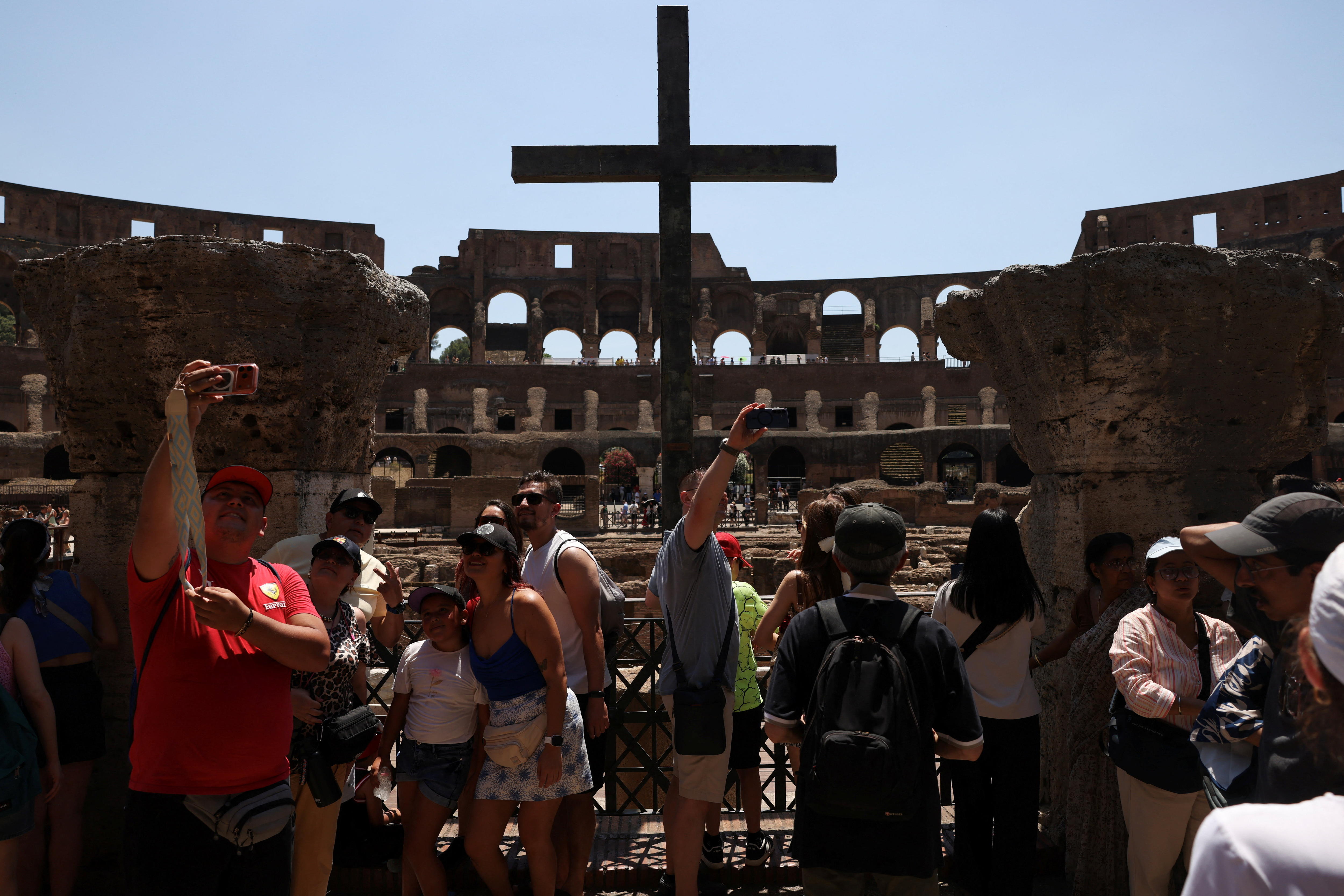 Tourists taking selfies in small groups on a tiered interior level of the Colosseum on a blue-sky day.
